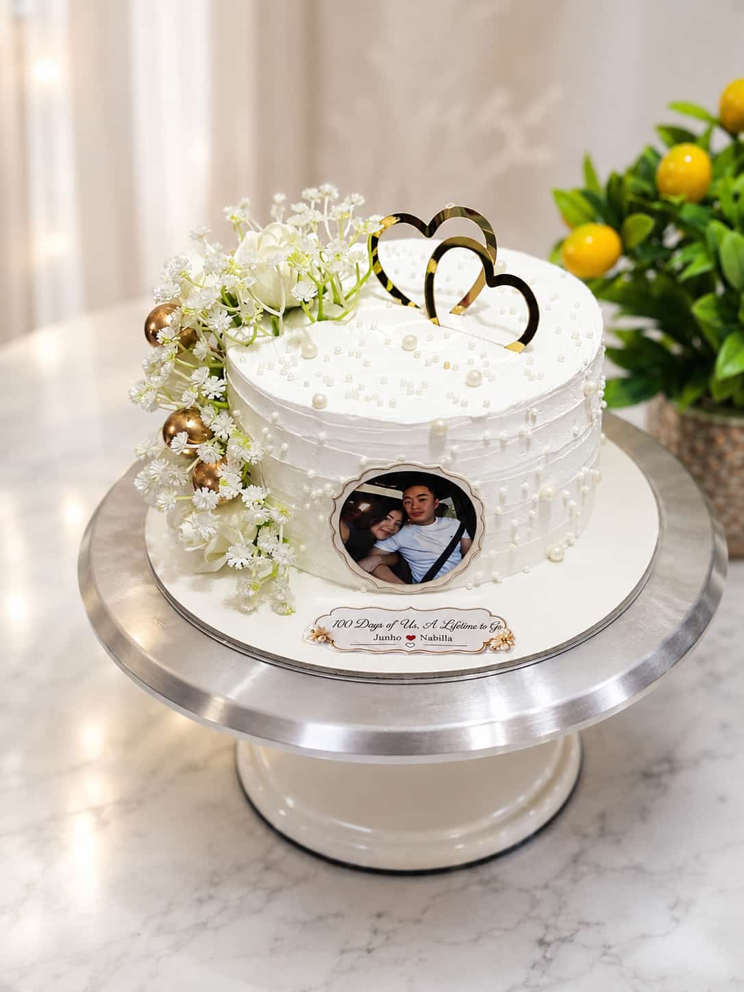 White textured buttercream anniversary cake with pearl details, gold hearts, fresh white flowers and a circular edible photo print of a couple, personalised '100 Days of Us', by CakeCanva in Bahrain.