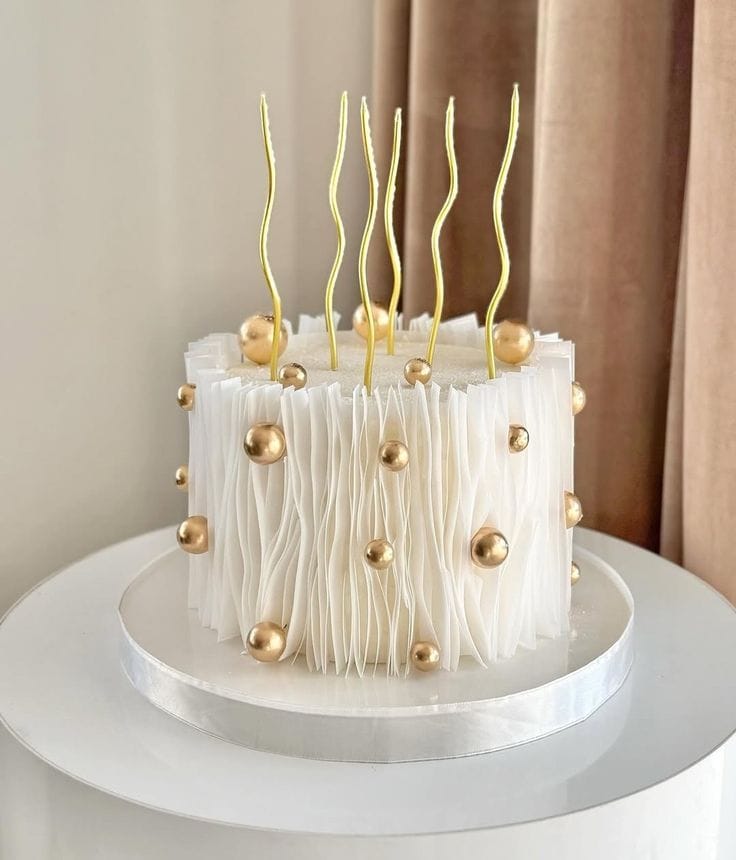 White pleated buttercream cake textured like fabric folds, decorated with gold pearls and gold squiggle candles, a signature anniversary cake style in Bahrain.