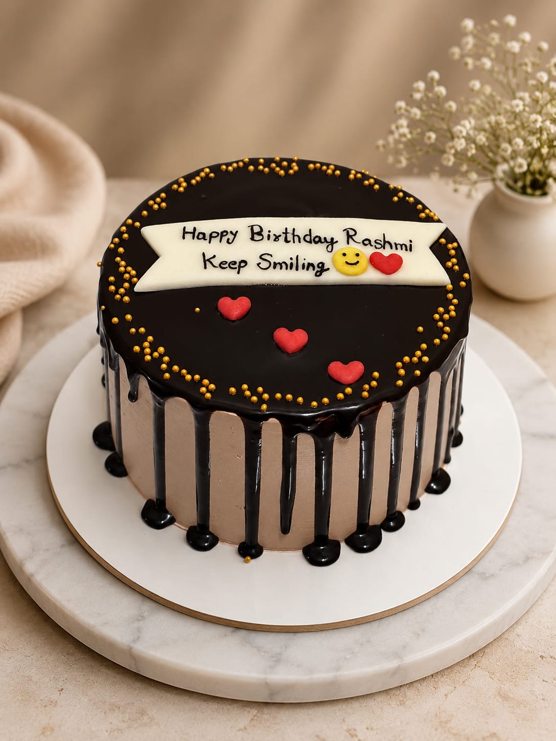 Chocolate cake with a glossy dark ganache mirror top and drip, gold sugar-pearl scatter, red fondant hearts and an ivory banner plaque reading 'Happy Birthday Rashmi Keep Smiling' in Bahrain.