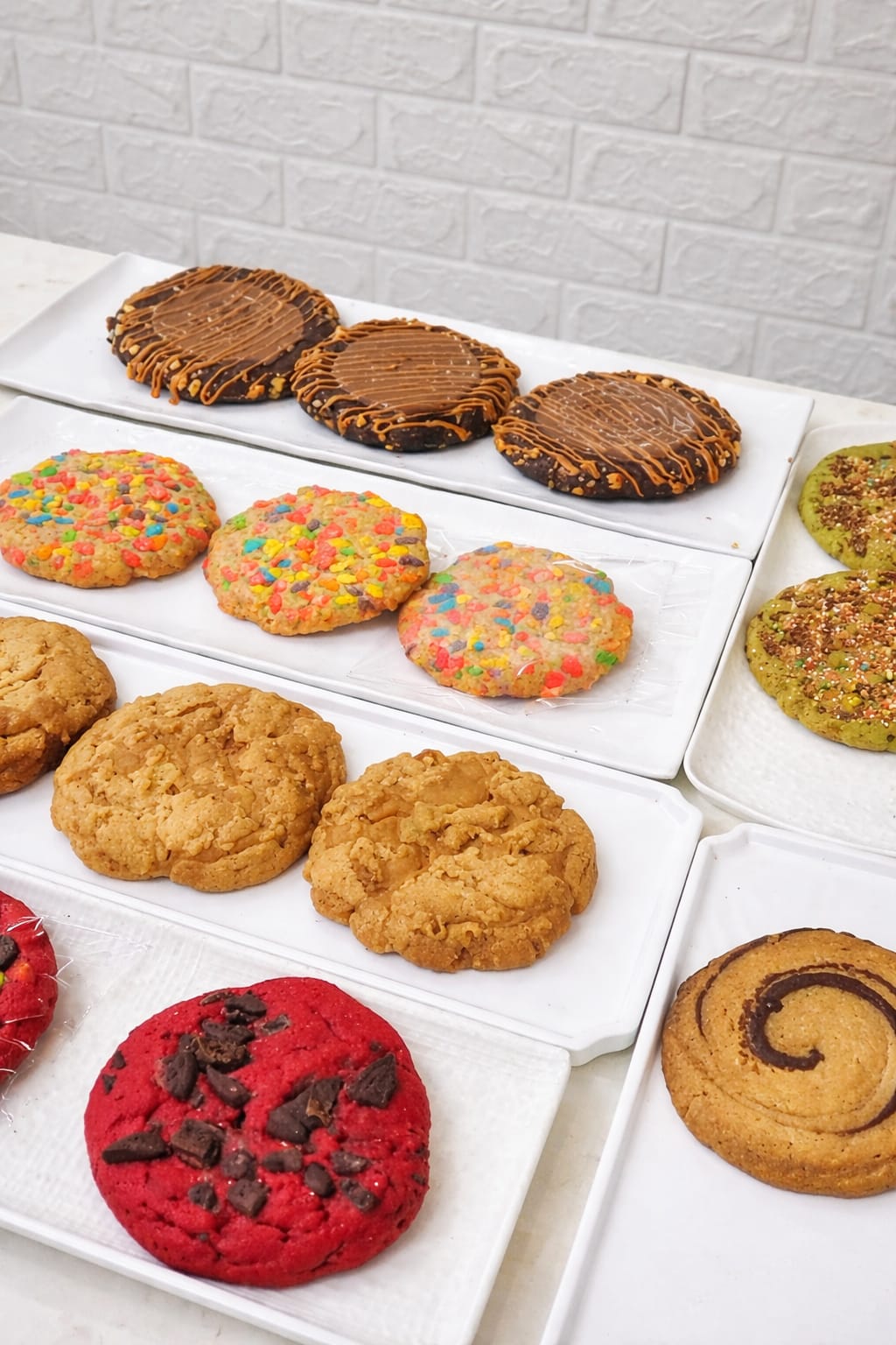Assorted gourmet cookies on white trays: chocolate with caramel drizzle, colourful cereal-topped, peanut butter, red velvet with chocolate chunks, pistachio and a cinnamon swirl cookie, by CakeCanva in Bahrain.