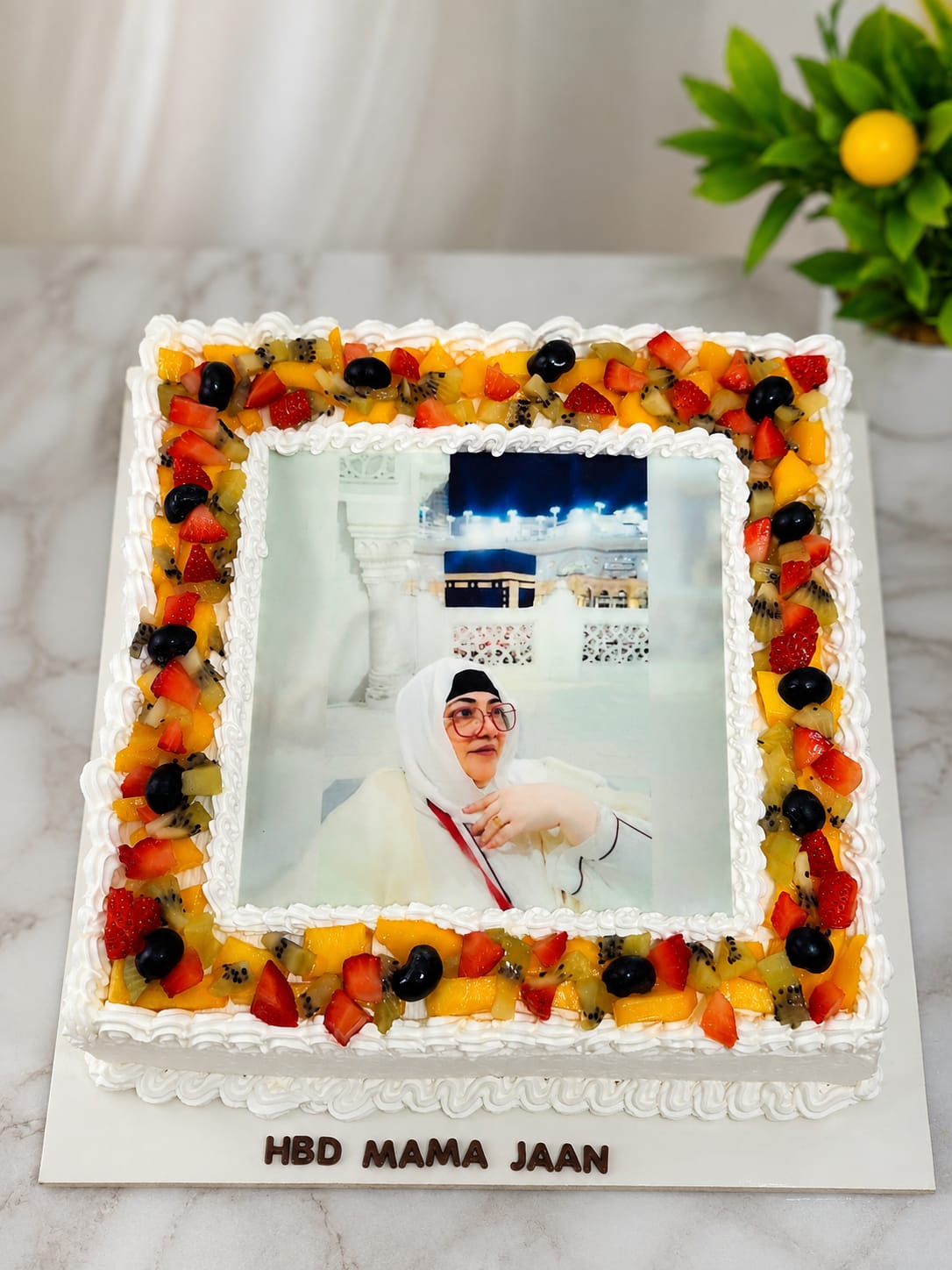 Square cake bordered with fresh strawberries, kiwi, mango and grapes framing an edible photo print of a woman at the Kaaba, personalized 'HBD Mama Jaan' in Bahrain.