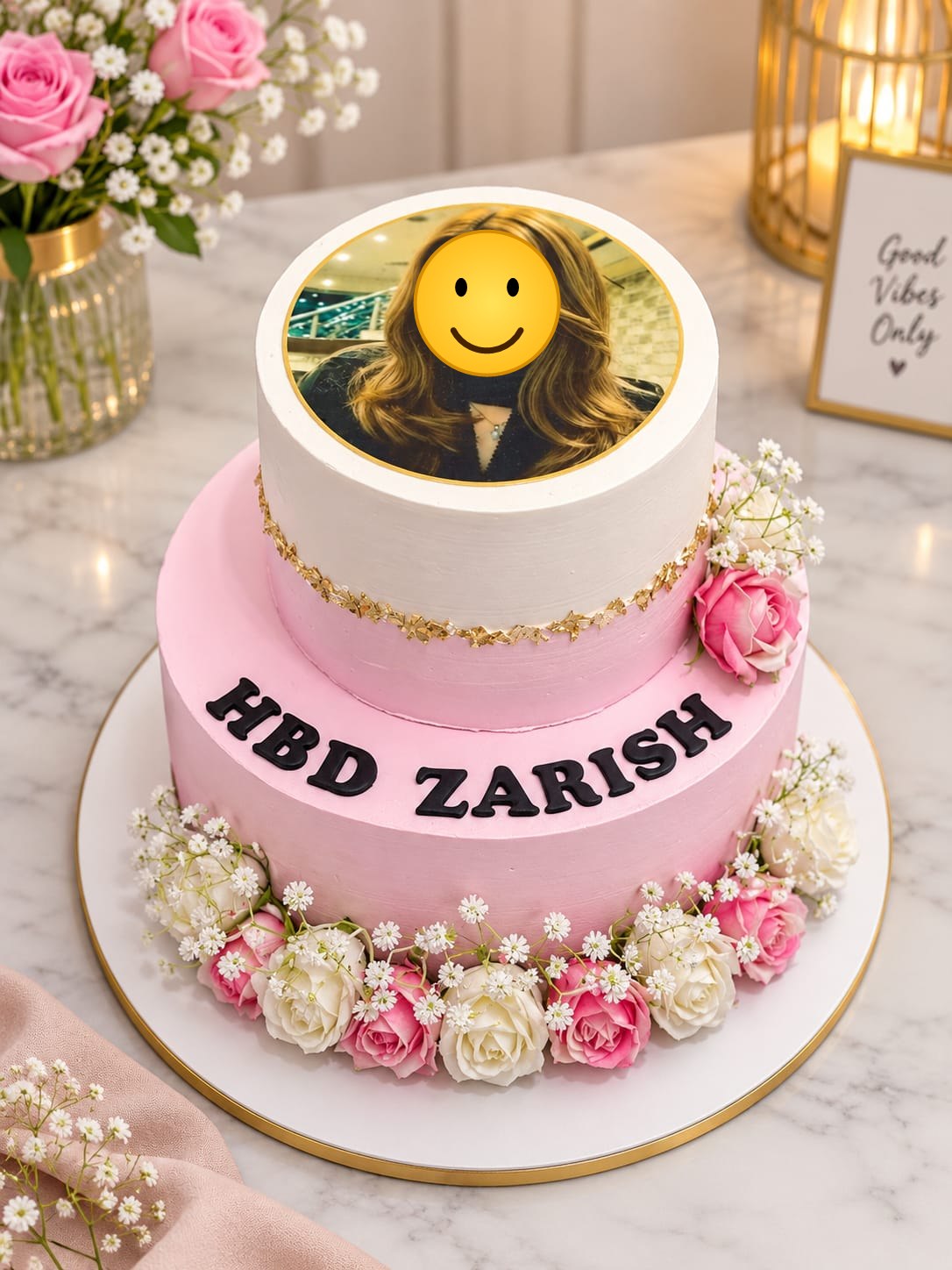 Two-tier pink and white buttercream cake with a round edible photo print on the top tier, a gold leaf band, fresh pink and white roses with baby's breath at the base and black fondant 'HBD Zarish' lettering, in Bahrain.