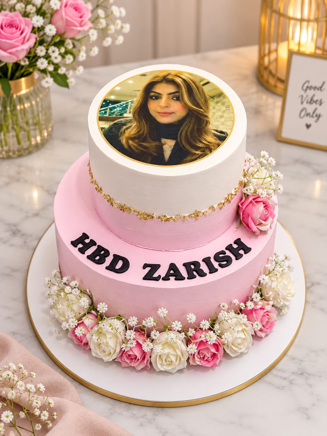 Two-tier pink and white buttercream cake with a round edible photo print on the top tier, a gold leaf band, fresh pink and white roses with baby's breath at the base and black fondant 'HBD Zarish' lettering, in Bahrain.