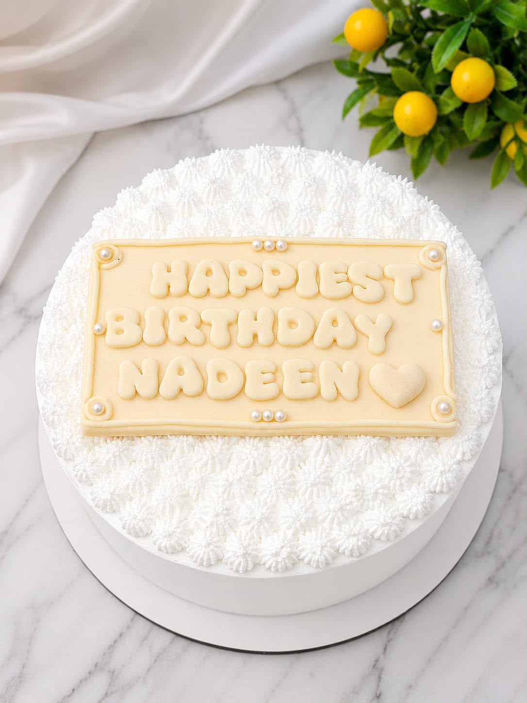 White cake with an all-over piped lace buttercream texture, topped with an ivory fondant plaque reading 'Happiest Birthday Nadeen' in bubble lettering with pearl accents, in Bahrain.