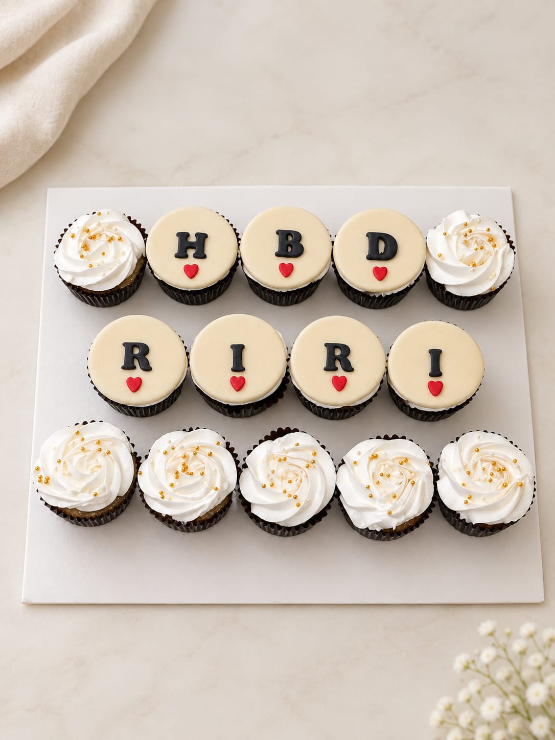 Set of fourteen cupcakes arranged to spell 'HBD RIRI' with ivory fondant discs, black lettering and red hearts, framed by white buttercream rosette cupcakes with gold sugar pearls, a personalised cupcake set in Bahrain.