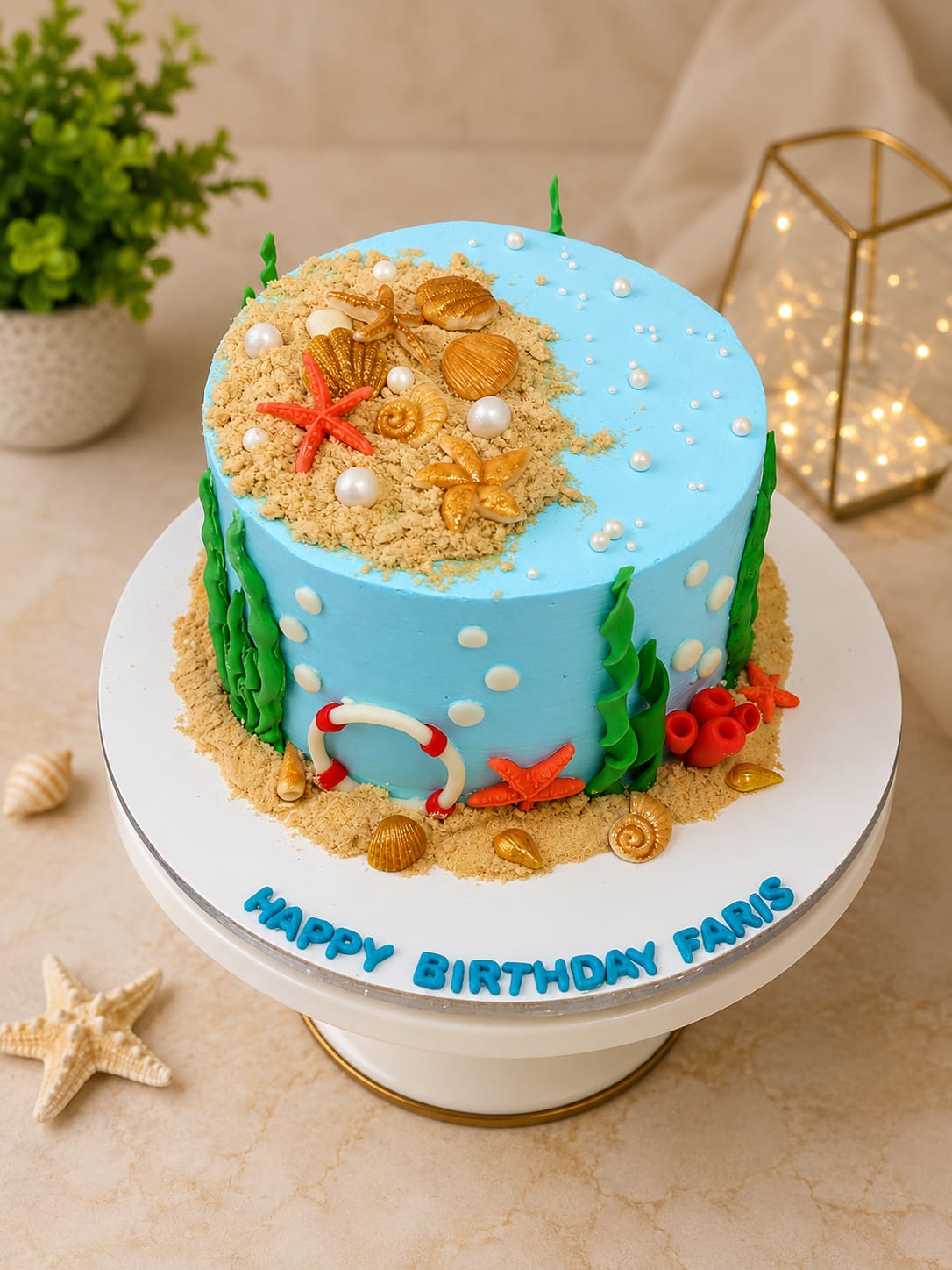 Blue buttercream under-the-sea theme birthday cake with a biscuit-crumb sandy beach, gold fondant seashells and starfish, green seaweed, sugar-pearl bubbles and a mini life ring, personalized 'Happy Birthday Faris' in Bahrain.