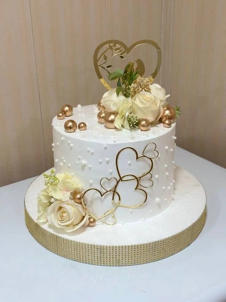 White textured buttercream wedding cake decorated with a gold wire heart topper, ivory roses, and gold pearls, an elegant wedding cake style seen in Bahrain.