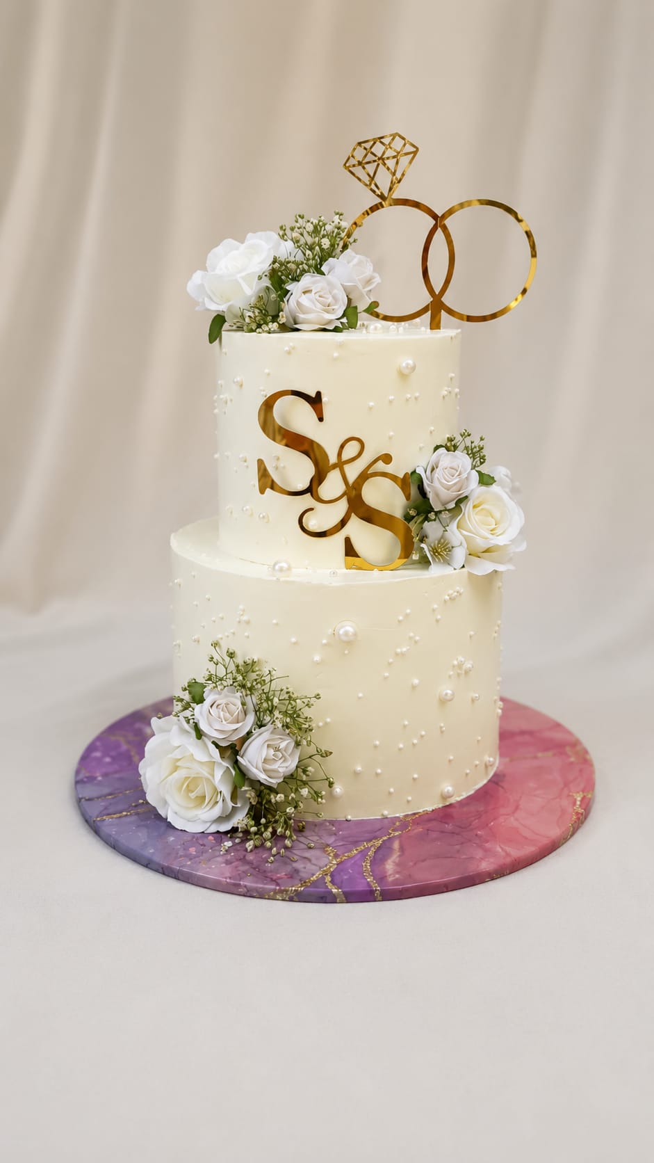 Two tier ivory wedding cake decorated with pearl details, fresh white rose flowers, and a gold monogram with a diamond ring topper, styled for a Bahrain wedding.